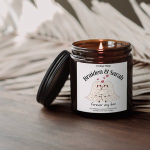 May include: A lit, brown glass jar candle with a black lid. The label reads "Braiden & Sarah forever my boo" with illustrations of two ghost figures and hearts. The candle is on a wooden surface.