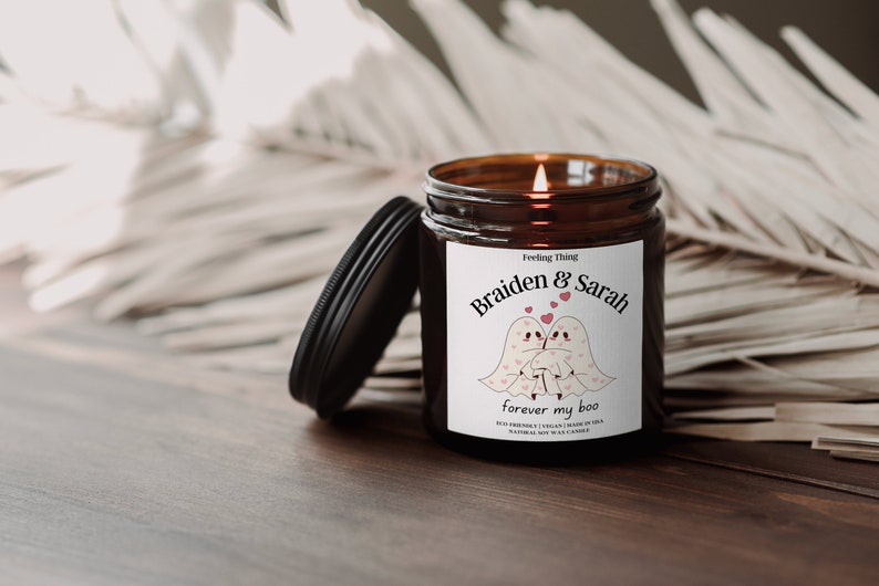 May include: A lit, brown glass jar candle with a black lid. The label reads "Braiden & Sarah forever my boo" with illustrations of two ghost figures and hearts. The candle is on a wooden surface.