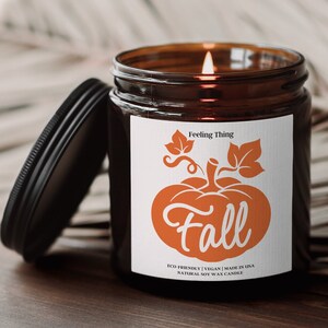 May include: A lit soy wax candle in a brown glass jar with a black lid. The label features an orange pumpkin graphic with the word "Fall". Text on the label reads "Feeling Thing" and "Eco-Friendly | Vegan | Made in USA".