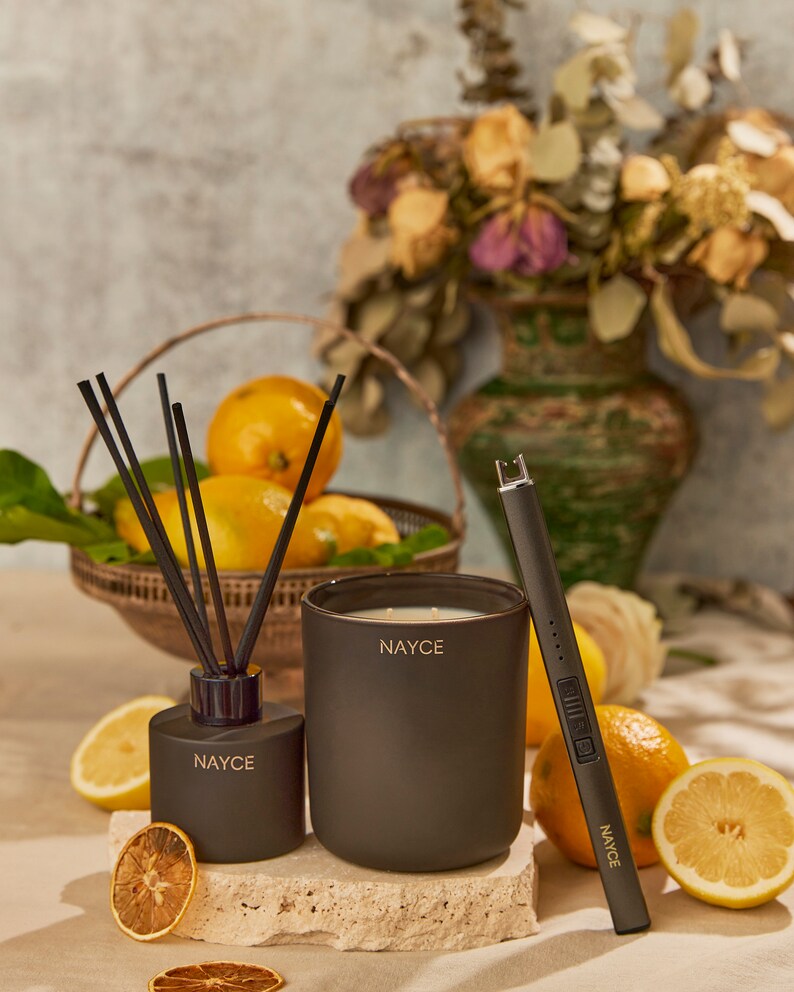 May include: A black candle, diffuser, and lighter with the word "NAYCE" printed on each. The candle and diffuser are on a stone surface with lemons and dried lemon slices around them.
