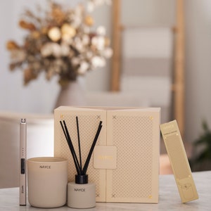 May include: A gift set for home fragrance lovers featuring a candle, diffuser, and a candle lighter. The set is packaged in a beige box with a geometric pattern and the brand name "NAYCE" printed on it. The candle and diffuser are both in a beige color with the brand name printed on them.