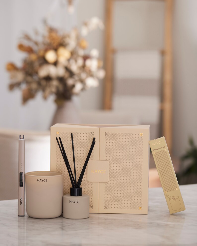 May include: A gift set for home fragrance lovers featuring a candle, diffuser, and a candle lighter. The set is packaged in a beige box with a geometric pattern and the brand name "NAYCE" printed on it. The candle and diffuser are both in a beige color with the brand name printed on them.