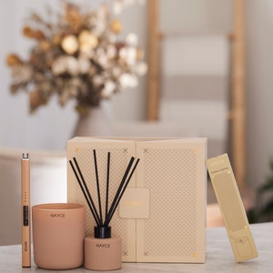 May include: A gift set featuring a beige candle, a diffuser with black reeds, and a gold candle lighter. The set is packaged in a beige box with a geometric pattern and the word "NAYCE" printed on it.
