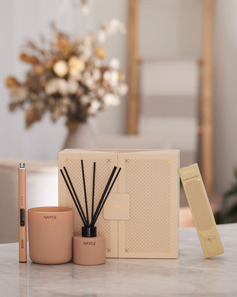 May include: A gift set featuring a beige candle, a diffuser with black reeds, and a gold candle lighter. The set is packaged in a beige box with a geometric pattern and the word "NAYCE" printed on it.