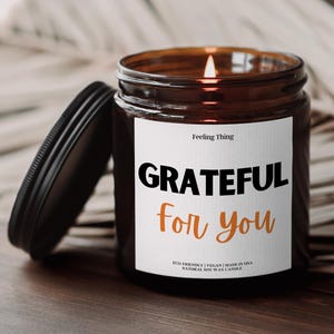 May include: A lit soy wax candle in a brown glass jar with a black lid. The label reads "GRATEFUL for you" in black and orange text. The candle is eco-friendly, vegan, and made in the USA.