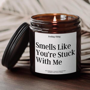 May include: A brown glass candle with a white label that reads "Smells Like You're Stuck With Me". The candle is on a wooden surface. The label also says "Feeling Thing", "Eco-Friendly", "Vegan", "Made in USA", and "Natural Soy Wax Candle".
