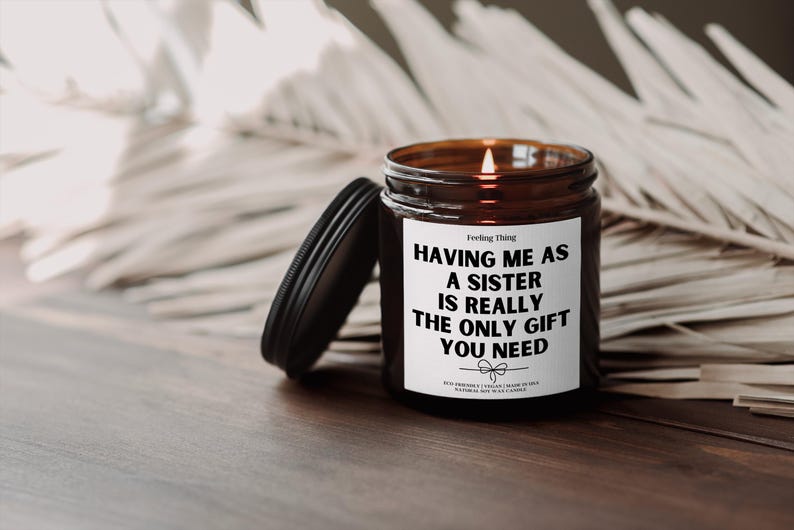 Op de afbeelding: Een bruine glazen kaars met een wit label waarop staat "Het hebben van een zus is echt het enige cadeau dat je nodig hebt". De kaars is aangestoken en de vlam is zichtbaar.