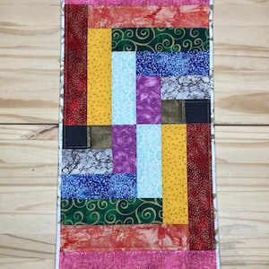 May include: A rectangular, handmade quilt with a patchwork design. The quilt features a variety of fabrics in shades of pink, red, yellow, green, blue, and purple. The design includes rectangular and square blocks, with a white border.