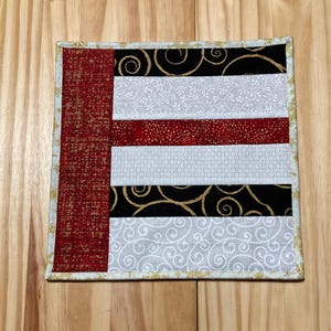 May include: A square quilted coaster with a patchwork design. The coaster features a red textured fabric on the left side and alternating horizontal stripes of black with gold swirls, white with a floral pattern, and red with gold dots. The edges are trimmed with a gold snowflake pattern.