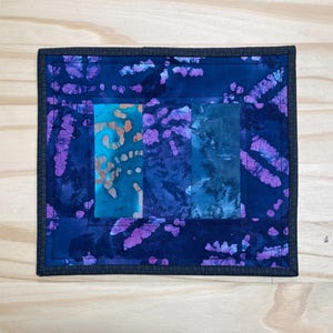May include: A square, handmade textile art piece with a dark blue border and a central design of vertical rectangles. The rectangles feature shades of blue, teal, and purple with batik-style patterns. The piece is likely a small quilt or placemat.