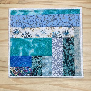 May include: A square quilted coaster featuring a patchwork design. The coaster showcases various blue fabrics, including swirling patterns, snowflakes, and tie-dye effects. It is bordered with a white fabric and a white stitched edge.