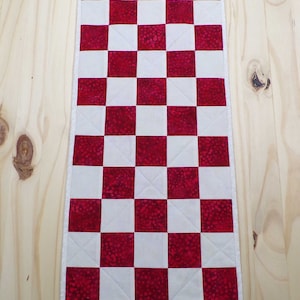 May include: A rectangular quilted table runner with a checkerboard pattern of red and white squares. The red squares have a mottled texture. The runner is bordered with a thin white edge and is displayed on a light wood surface.
