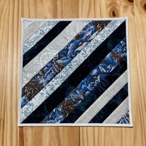May include: A square quilted table runner with a white border and a diagonal pattern of black, white, and blue fabric strips. The fabric strips feature a winter scene with snow-covered trees and houses.