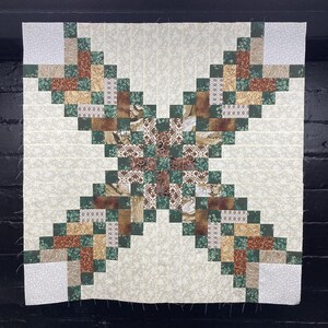May include: A quilt block with a green, brown, and white geometric pattern. The block is made up of small squares and is set against a cream background.