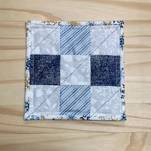 May include: Square quilted coaster with a patchwork design. The coaster features nine squares in shades of blue and white, with floral and striped patterns. The border is white with gold and blue floral accents.