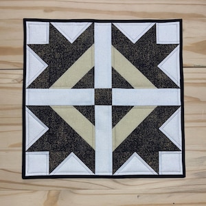 May include: Square quilted textile with a geometric design. The design features black, gold, white, and beige shapes arranged in a symmetrical pattern. The edges are finished with a black border. The textile is placed on a light wood surface.