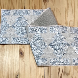 May include: A blue and white quilted table runner with a geometric pattern. The runner is folded in half and is lying on a wooden surface.
