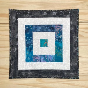 May include: Square quilted wall hanging with a log cabin design. The center is a square of teal fabric, surrounded by white, teal, and patterned fabrics. The border is a dark gray fabric with a mottled pattern.