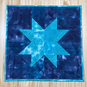 May include: A blue and teal patchwork quilt with a star design. The quilt is made of squares of fabric sewn together to create a star shape in the center. The quilt is edged with a light blue fabric.