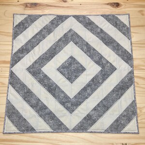 May include: A square quilt with a geometric design. The quilt features alternating bands of light gray and off-white fabric, creating a diamond pattern. The edges are finished with a gray binding. The quilt is displayed on a wooden surface.