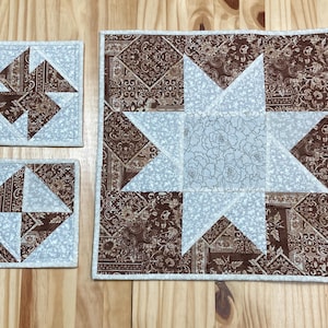 May include: Three quilted coasters with a brown and white star pattern. The coasters are made of fabric and have a white border.