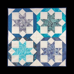 May include: A square quilt with a star pattern in shades of blue and white. The quilt features four large star designs, each with a different patterned fabric. The stars are set against a white background with small black dots, and the quilt is bordered with a dark blue binding.