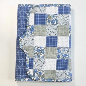 May include: A quilted notebook cover in shades of blue, white, and gray. The cover features a patchwork design with squares of various patterns, including floral prints and grid patterns. The edges are finished with a scalloped trim.