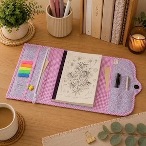 Op de afbeelding: Een stoffen notitieboekhoes met een roze en wit gingham exterieur en een bloemrijke voering. De hoes bevat interne vakken voor pennen, een notitieboek met een botanische illustratie op de kaft en kleurrijke zelfklevende notitieblaadjes.