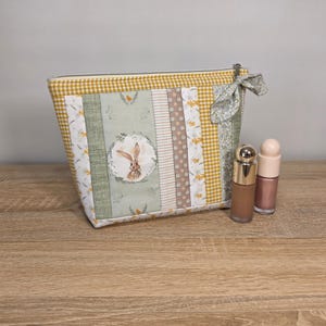 Peut inclure: Une trousse de maquillage patchwork avec une illustration de lapin, avec un motif vichy jaune et des accents floraux. Deux flacons de maquillage sont à côté du sac. Le sac a une fermeture éclair et un nœud décoratif.