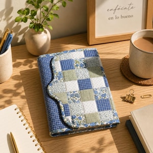 Könnte beinhalten: Ein gesteppter Notizbuchumschlag mit einem Patchwork-Design in Blau-, Grün- und Weißtönen. Der Umschlag hat einen gewellten Rand und ist offen. Im Hintergrund ist der Text "enfócate en lo bueno" in einem gerahmten Bild zu sehen.