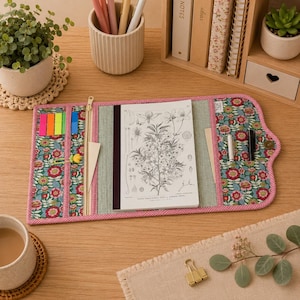 Puede incluir: Una funda de tela para cuaderno con estampado floral en tonos rojos, verdes y azules, con un borde de cuadros vichy rosa. El interior incluye un bolsillo con cremallera, varias ranuras para bolígrafos y notas adhesivas, y una funda central.