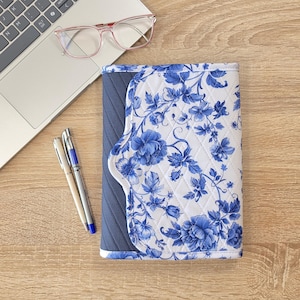 May include: A quilted notebook cover with a white background and blue floral pattern. The cover has a navy blue spine and a curved edge. A pair of pens, a laptop, and pink glasses are also visible.