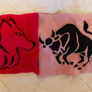 May include: Two fabric squares with embroidered animal designs. The left square is red with a black wolf silhouette. The right square is pink with a black bull silhouette.