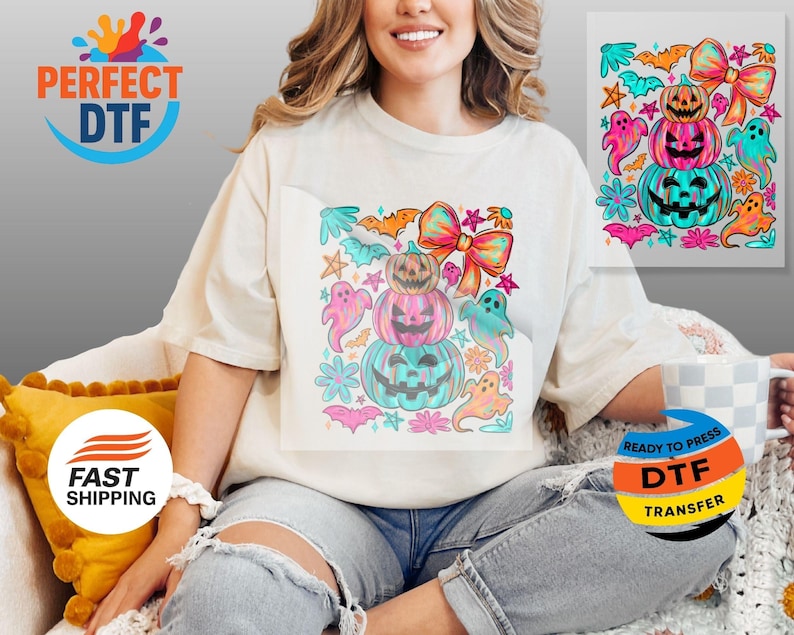 May include: A cream-colored t-shirt featuring a colorful Halloween-themed graphic. The design includes stacked pumpkins, ghosts, bats, and bows in shades of pink, teal, orange, and purple. The shirt has a relaxed fit and is suitable for the fall season.