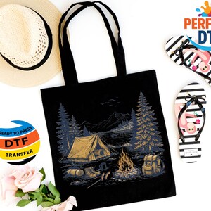 May include: Black tote bag with a camping scene graphic, a straw hat, and black and white striped flip-flops with flamingo designs. The tote bag features a tent, campfire, and forest illustration. A DTF transfer sticker is also visible.