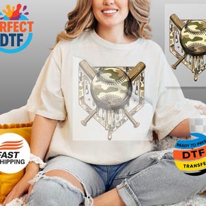May include: Cream-coloured t-shirt featuring a baseball design with a camouflage pattern. The design includes a baseball, crossed bats, and a home plate shape with dripping details. The shirt is a relaxed fit.