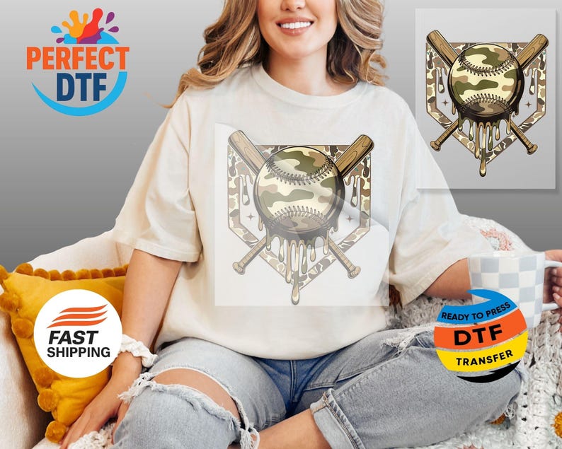 May include: Cream-coloured t-shirt featuring a baseball design with a camouflage pattern. The design includes a baseball, crossed bats, and a home plate shape with dripping details. The shirt is a relaxed fit.