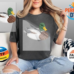 May include: A dark gray t-shirt featuring a duck design. The duck has a green plaid head, a yellow beak, and a brown patterned body. The duck is set against a white background.