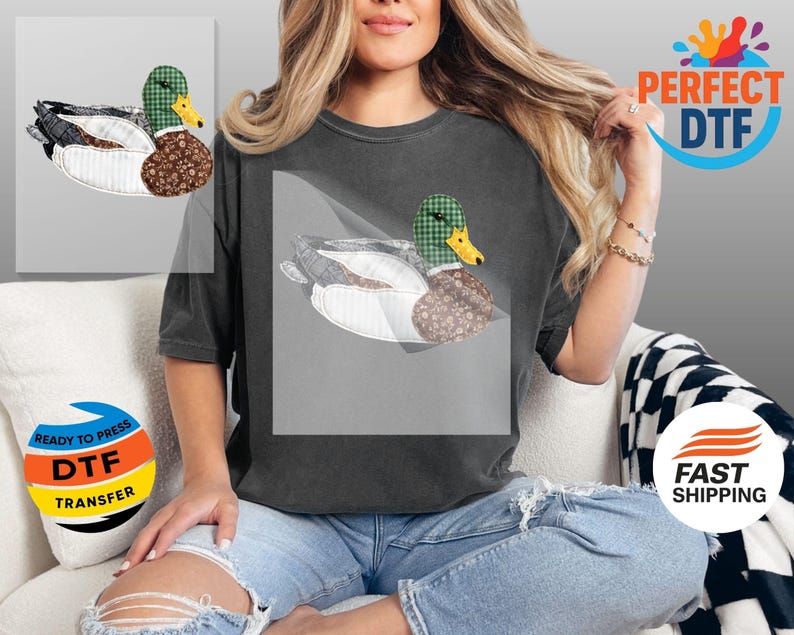 May include: A dark gray t-shirt featuring a duck design. The duck has a green plaid head, a yellow beak, and a brown patterned body. The duck is set against a white background.