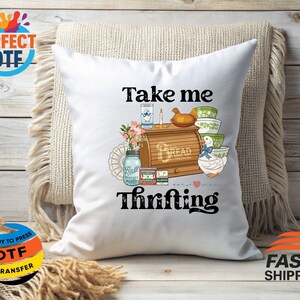 May include: White cushion with the words "Take me Thrifting" in black script. The cushion features a colourful illustration of a bread box, bowls, a jar, and a duck. The cushion is on a beige fringed cushion.