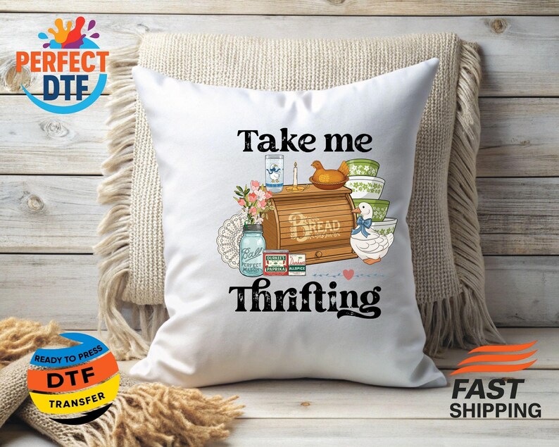 May include: White cushion with the words "Take me Thrifting" in black script. The cushion features a colourful illustration of a bread box, bowls, a jar, and a duck. The cushion is on a beige fringed cushion.
