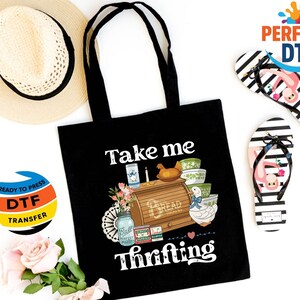 May include: Black tote bag with the text "Take me thrifting" and a graphic of a bread box, bowls, and a duck. A straw hat and black and white striped flip-flops with a flamingo design are also visible.