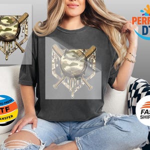 May include: A dark grey t-shirt with a baseball design. The design features a baseball with a camouflage pattern, crossed bats, and a dripping effect. The words "READY TO PRESS DTF TRANSFER" are also visible.