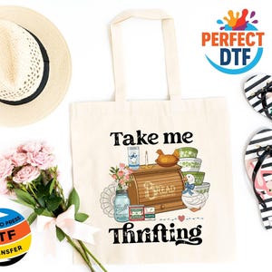 May include: A cream tote bag with the words "Take me Thrifting" and a graphic of vintage kitchen items. A straw hat, pink flowers, and black and white striped flip-flops with flamingo designs are also in the image.