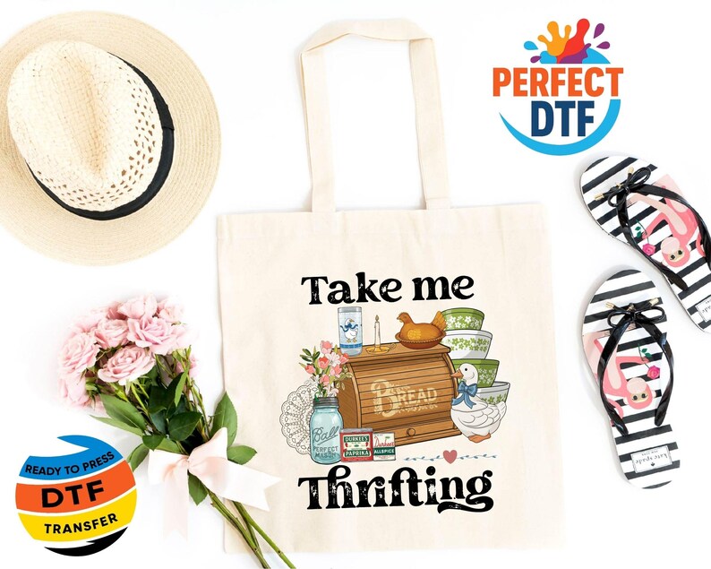 May include: A cream tote bag with the words "Take me Thrifting" and a graphic of vintage kitchen items. A straw hat, pink flowers, and black and white striped flip-flops with flamingo designs are also in the image.