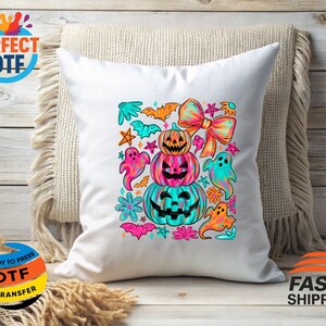 May include: White square pillow with a colorful Halloween design. The design features stacked pumpkins, ghosts, bats, and a large bow. The pumpkins are orange, pink, and teal. The ghosts are pink, teal, and orange. The pillow is on a beige throw.