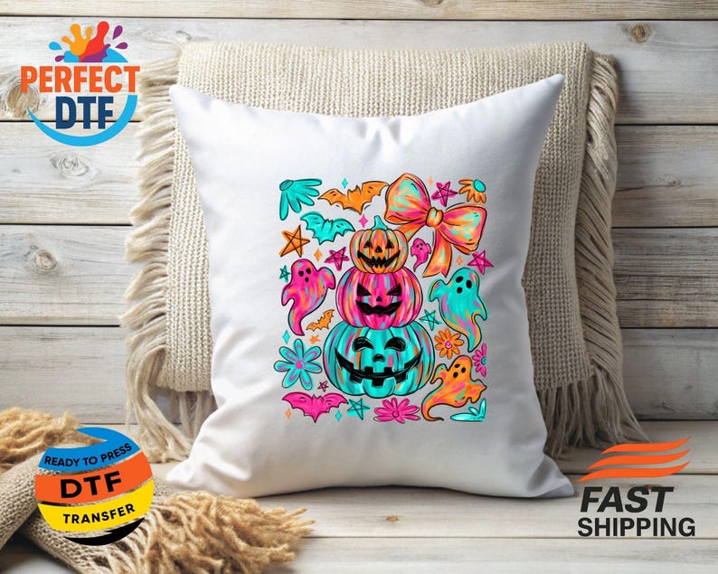 May include: White square pillow with a colorful Halloween design. The design features stacked pumpkins, ghosts, bats, and a large bow. The pumpkins are orange, pink, and teal. The ghosts are pink, teal, and orange. The pillow is on a beige throw.