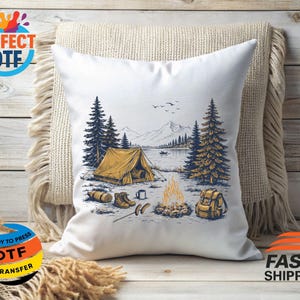 May include: White throw pillow with a camping scene illustration. The design features a yellow tent, campfire, lake, mountains, and trees. Additional elements include a backpack, boots, and a log. The pillow is resting on a beige blanket.