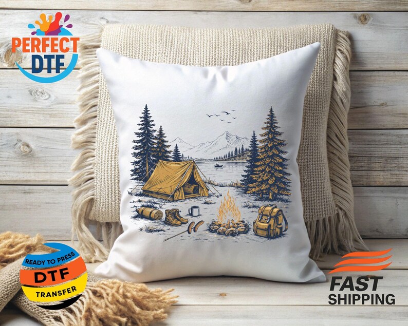 May include: White throw pillow with a camping scene illustration. The design features a yellow tent, campfire, lake, mountains, and trees. Additional elements include a backpack, boots, and a log. The pillow is resting on a beige blanket.