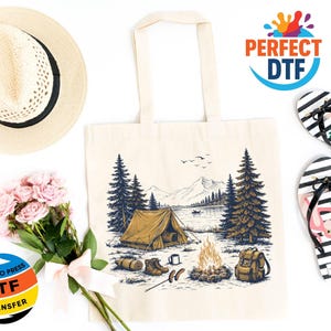 May include: Cream-colored tote bag with a camping scene illustration, featuring a tent, campfire, and mountains. The image includes a straw hat, pink flowers, and black and white striped flip-flops with a flamingo design. The text "READY TO PRESS DTF TRANSFER" is visible.